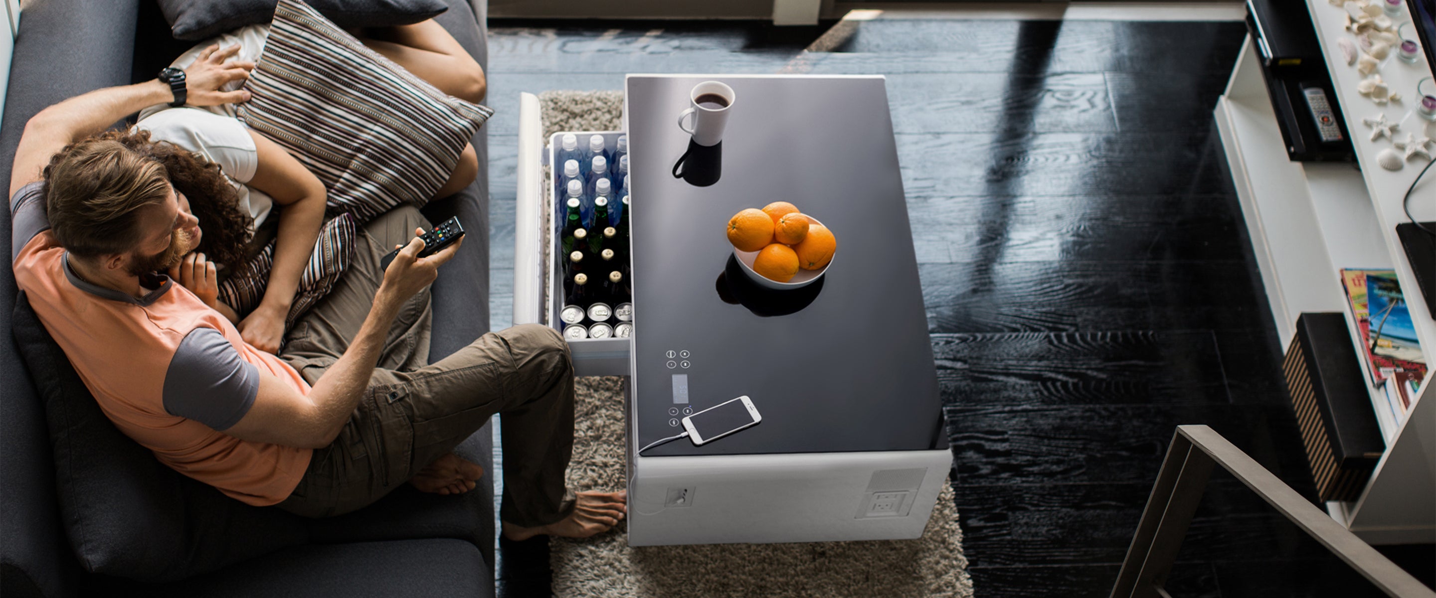 Man and woman relaxing on a sofa in a modern living room, with a Sobro smart coffee table in front of them featuring a built-in beverage cooler, and a smartphone charging.