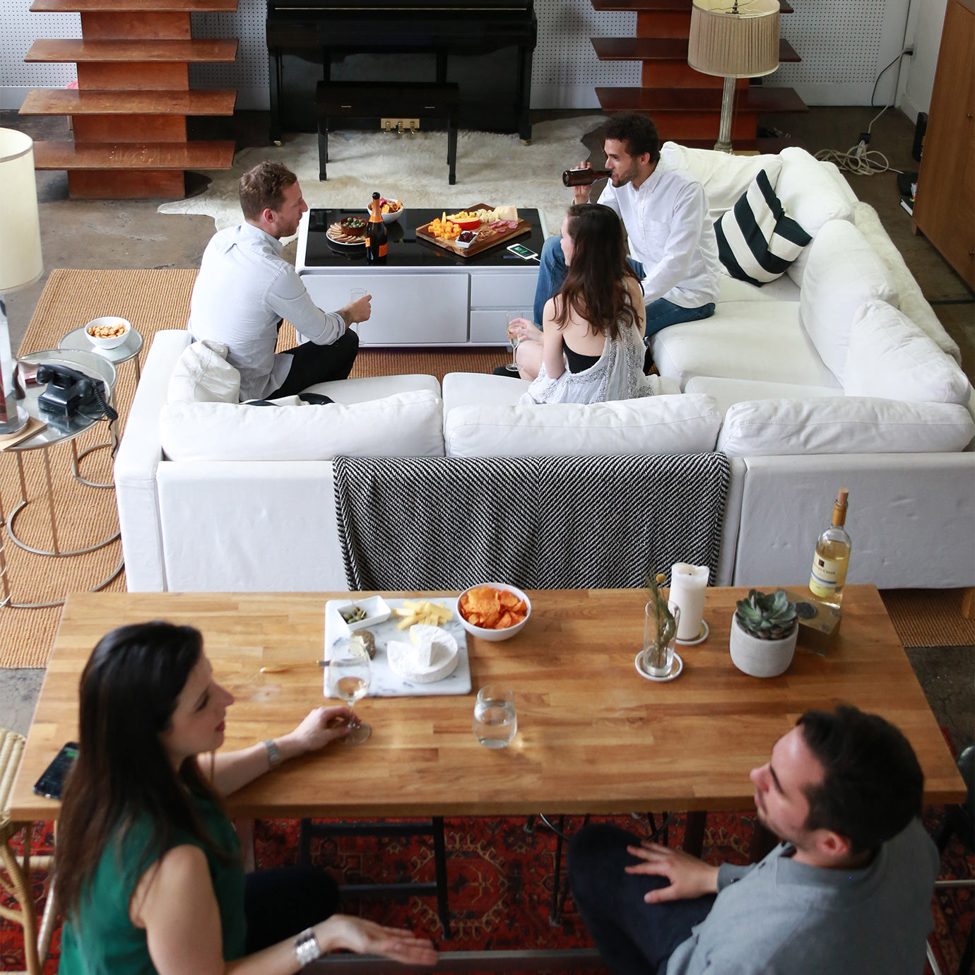 People gathered in a modern living room, chatting around a white sectional and smart coffee table with snacks.