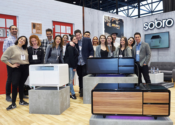 Sobro team gathered at a trade show booth, smiling in front of a display of smart coffee tables in various finishes.